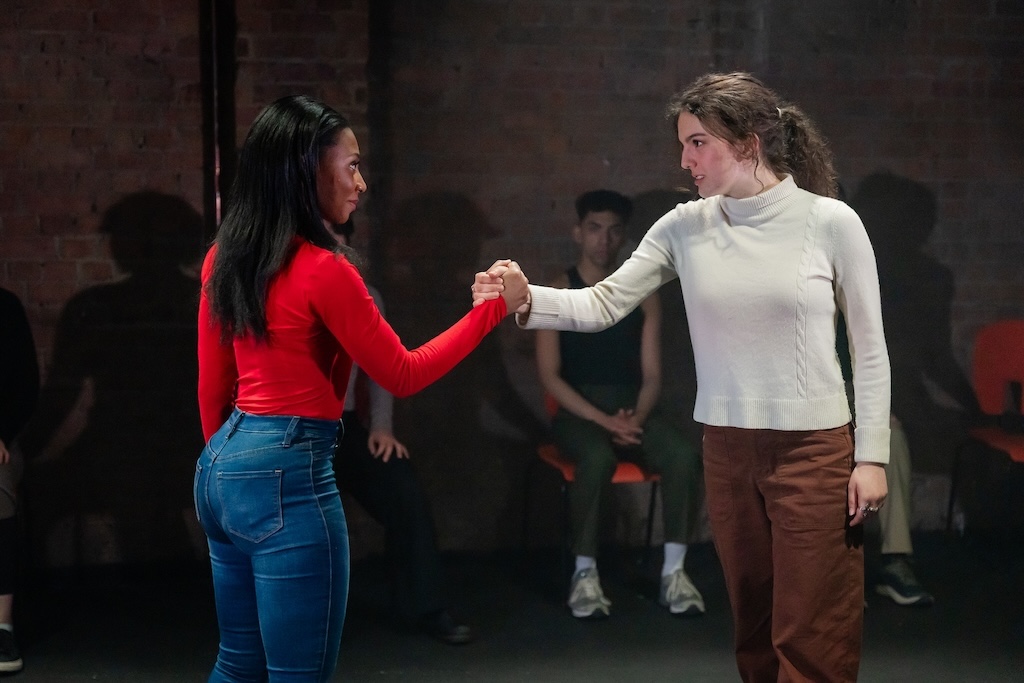 Action shot of the spring 2024 Shakespeare in Performance at RADA final presentation of Julius Caesar. Two actors look at each other and grasp hands on stage in mid-performance while other actors, seated behind them, look on.